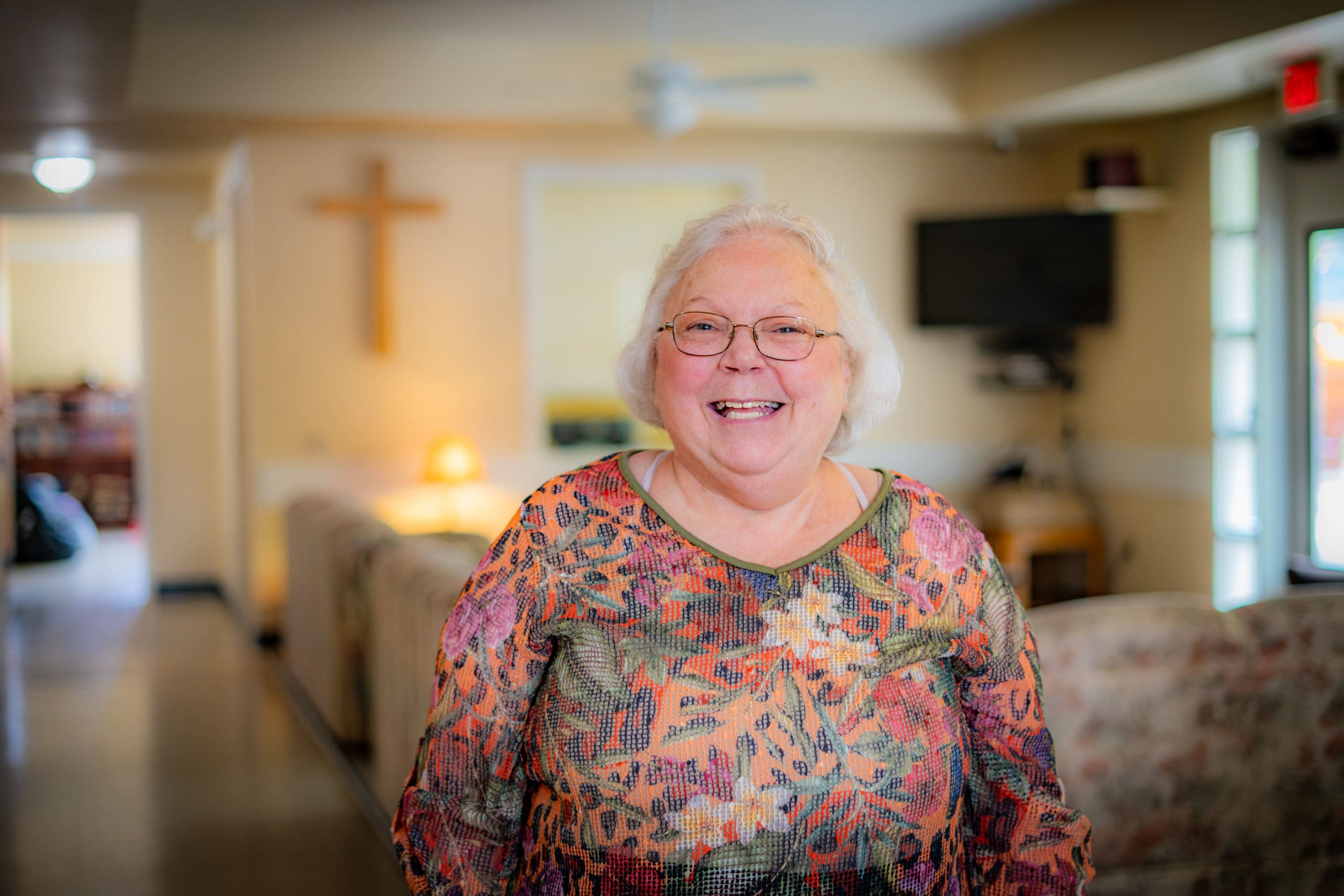 Margie stands smiling in the living room of Fikso Family Center, a cross can be seen in the background.