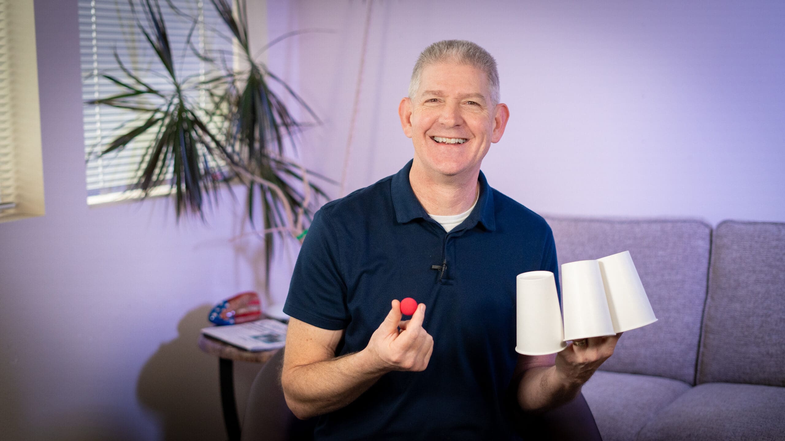 Brian holds 3 white paper cups, and one small, red ball.
