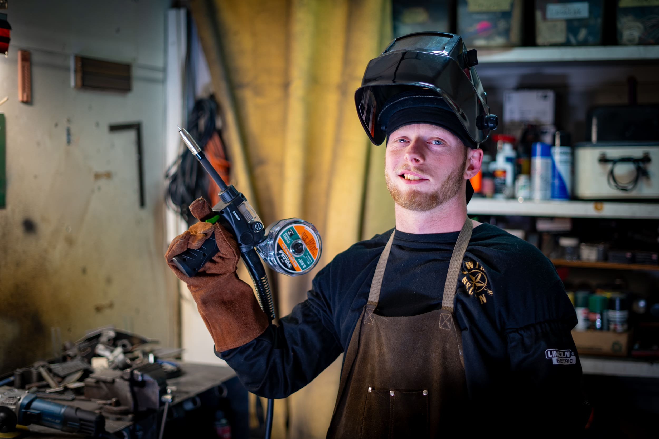 Brien holds a welding tool, wearing a welding mask.