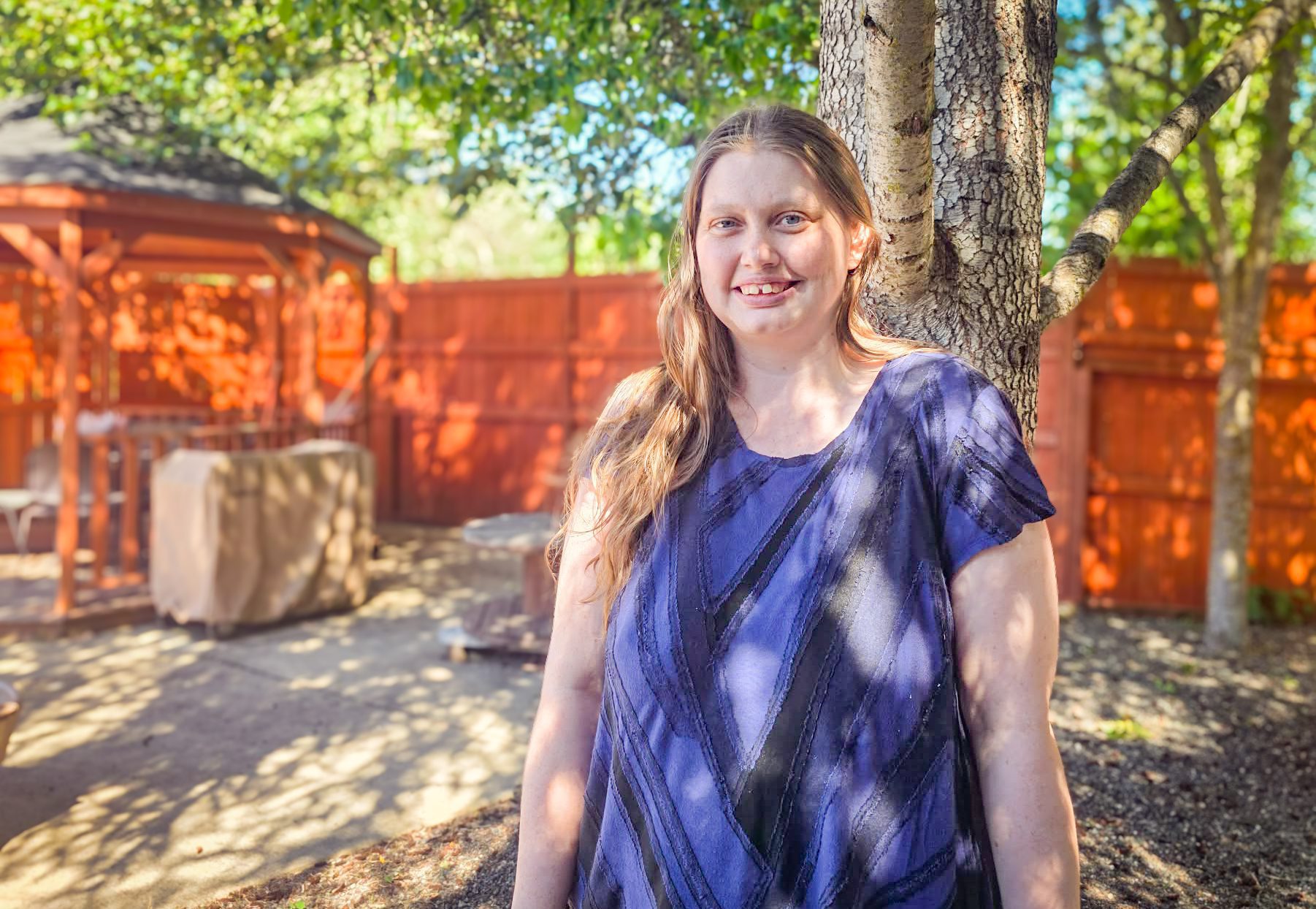 Smiling woman in a blue-purple blouse leaning against a tree in a sunny backyard with a wooden fence.