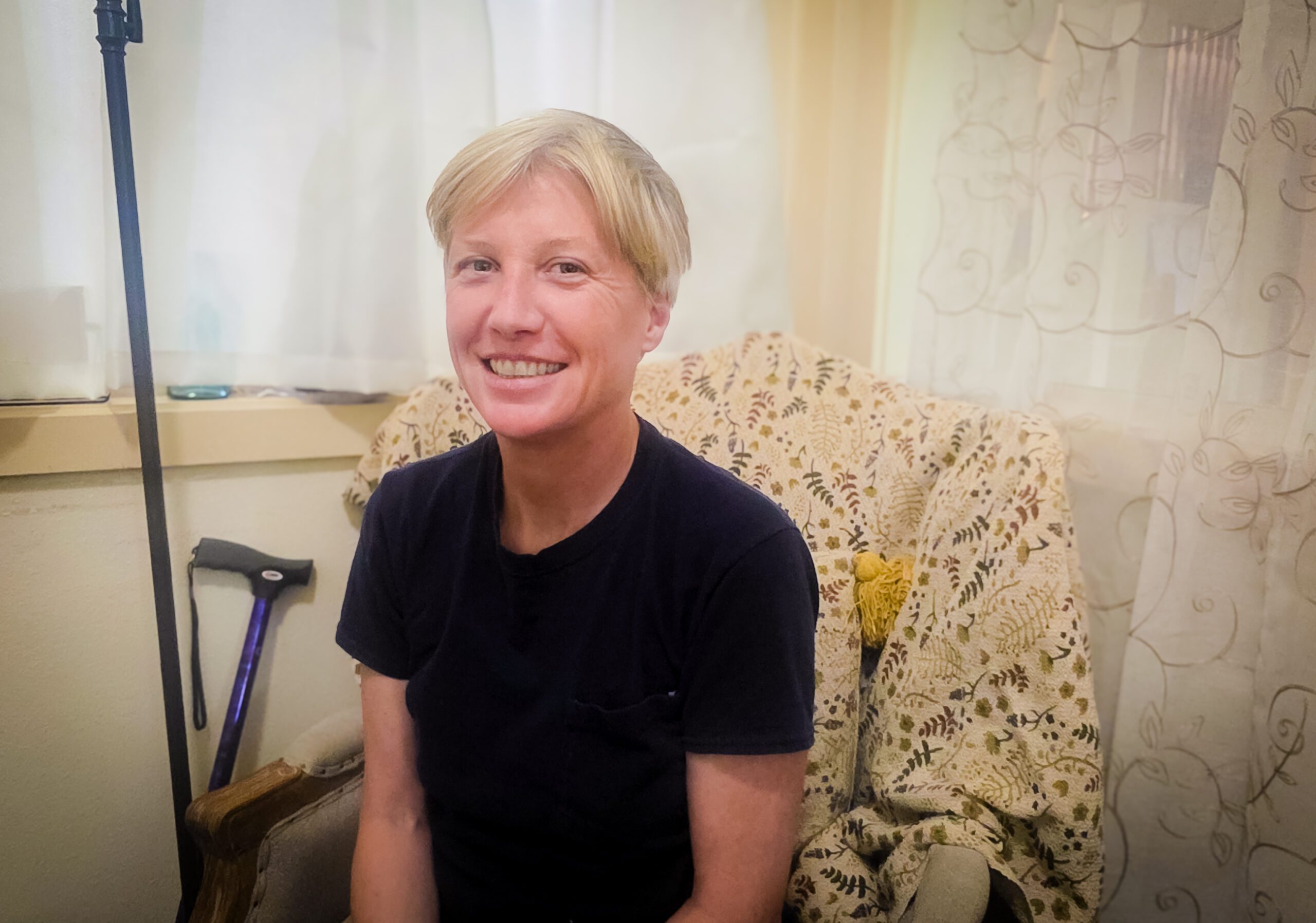 Smiling woman with short blonde hair in a black shirt, seated in a floral-patterned chair indoors.