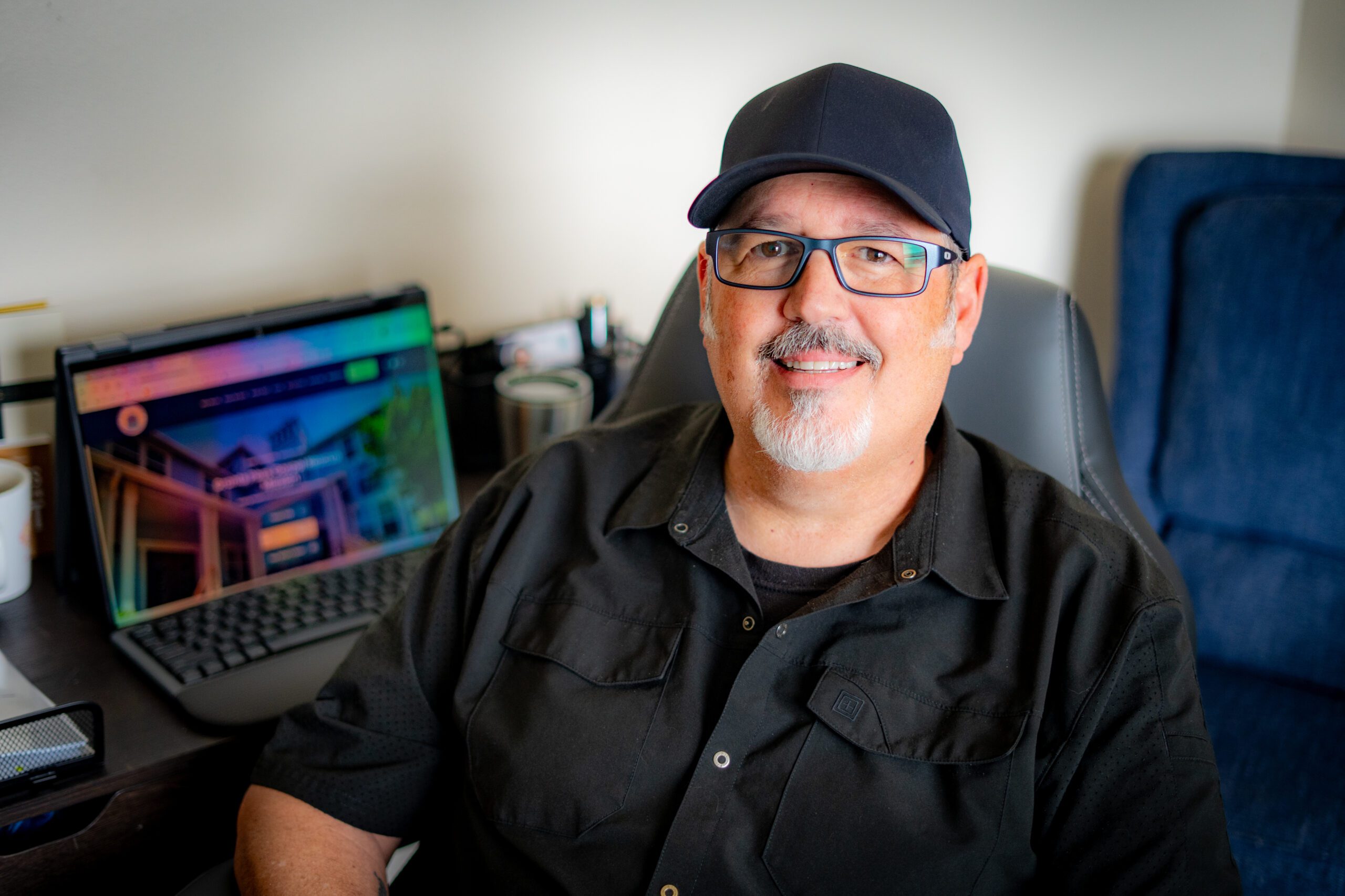 Smiling man wearing a dark cap and glasses seated at a desk with a computer monitor behind him in a home office setting.