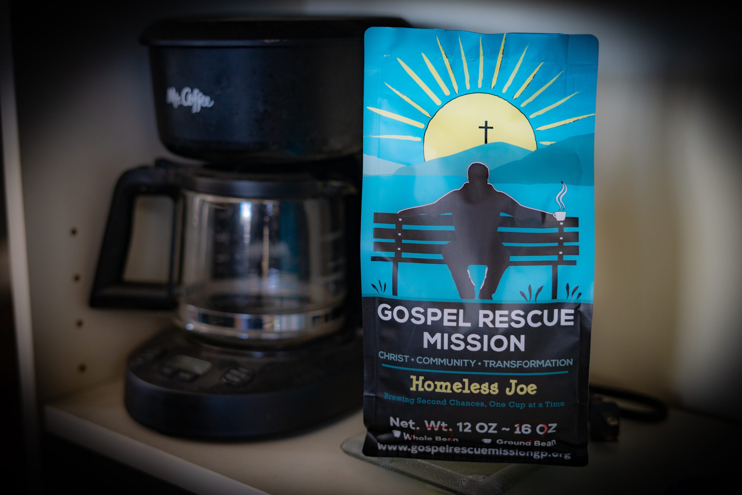 A blue Gospel Rescue Mission coffee bag on a kitchen shelf beside a coffee maker, featuring a sun, a bench, and the words 'Gospel Rescue Mission'.